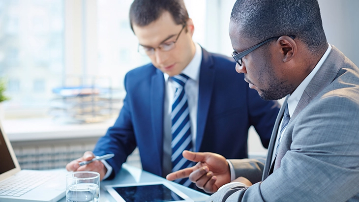 Two men sitting at desk looking at tablet device and talking