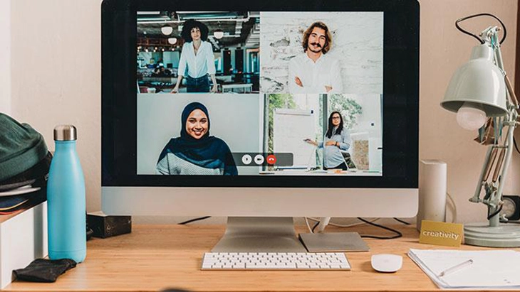 Large computer screen on desk displaying video call images of four people