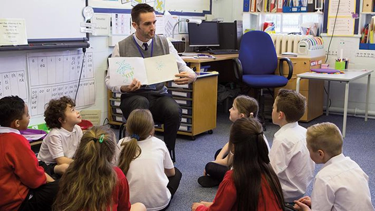 Teacher sitting in chair reading a book to young primary school students sitting on floor