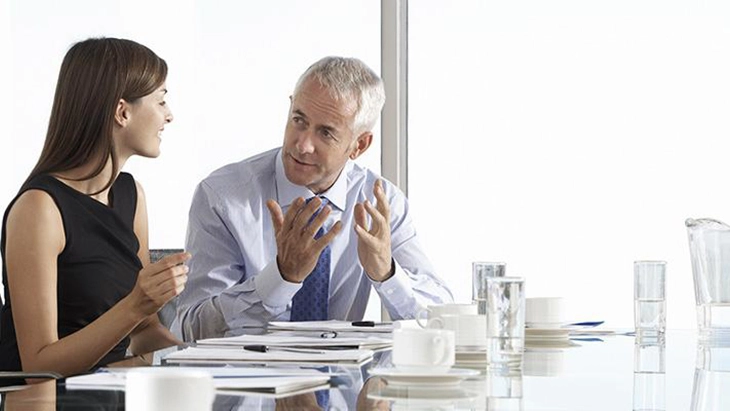 Man sitting at table in work space speaking to younger female colleague