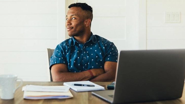 Man sitting in front of open lap top with arms crossed and a confident smile