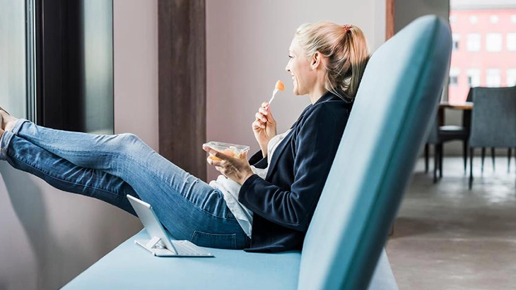 Young woman sitting on sofa, feet on window sill, eating lunch