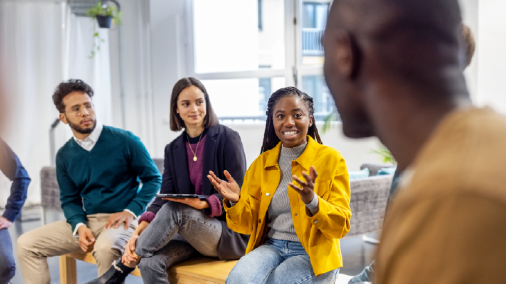 Group discussion in a modern office setting, with a person in a yellow jacket speaking while others listen attentively, representing teamwork and leadership development.